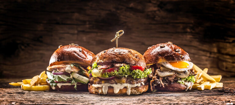 Drei große, reichhaltig belegte Burger auf einem rustikalen Holztisch. Von links nach rechts: ein Burger mit Lachs und frischem Gemüse, ein klassischer Burger mit Salat, Tomate und Käse, sowie ein Burger mit saftigem Rindfleisch, Pilzen und einem Spiegelei. Neben den Burgern sind goldgelbe Pommes Frites zu sehen. Alle Burger sind auf knusprigen, dunklen Brötchen serviert und mit frischen Zutaten belegt.