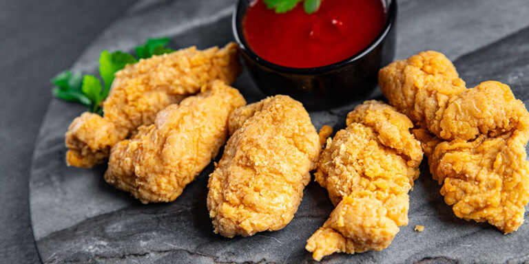 Knusprige, goldbraune Hähnchen-Nuggets auf einem dunklen Steinteller, begleitet von einer Schale mit roter Dip-Sauce. Die Nuggets sind appetitlich und gleichmäßig paniert. Im Hintergrund ist etwas grüne Petersilie zur Dekoration zu sehen