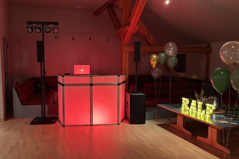 DJ-Setup mit beleuchteter, rot angestrahlter DJ-Konsole in einem festlich dekorierten Raum. Links stehen Lautsprecher und Lichttechnik, rechts bunte Luftballons und ein Tisch mit einem beleuchteten Schriftzug 'RALF' sowie floralen Dekorationen.